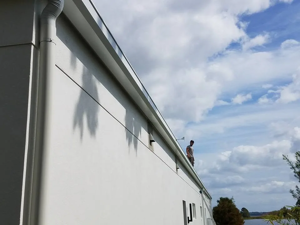Worker on roof edge long white stucco wall round downspout lakefront, Sanford
